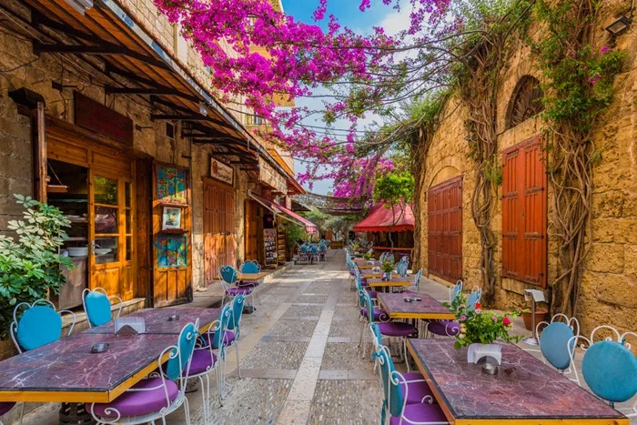 Cafe tables lining a stone-paved lane in the old souk of Byblos, Lebanon, shaded by pink bougainvillea, a charming stop on any travel itinerary