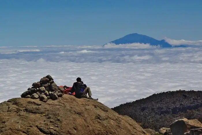 Climber taking a break on a rocky ridge on Kilimanjaro, looking out over the clouds toward Mount Meru