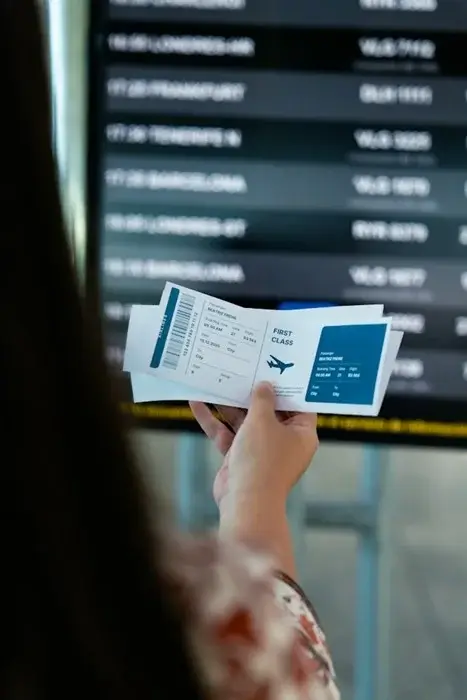 Traveler holding printed first-class flight tickets in front of an airport departures board, checking her onward ticket details before a visa review