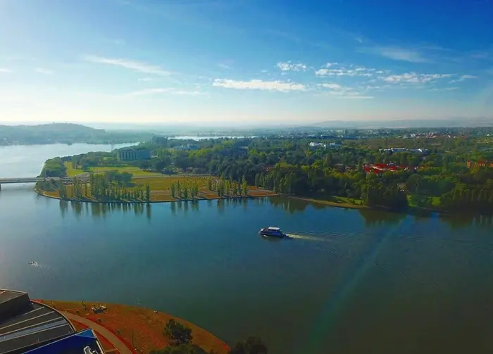 Aerial view of a cruise boat gliding across Lake Burley Griffin past leafy parkland, showing the green side of Canberra that first-time travelers love