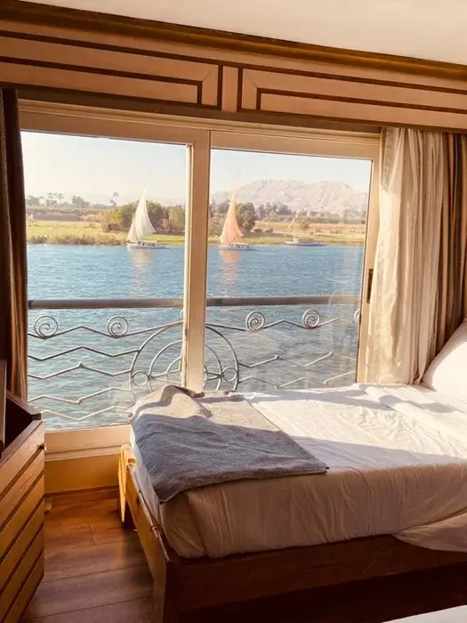 Cozy cruise cabin with a single bed and a large window framing feluccas sailing on the Nile