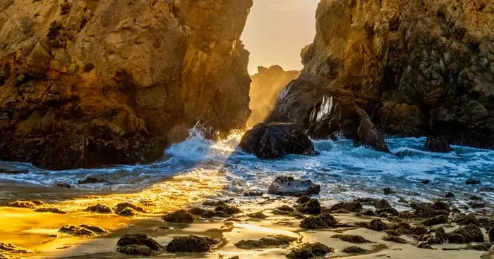 Golden sunset light streaming between sea stacks at Pfeiffer Beach in Big Sur, as waves wash over the rocky shore during golden hour