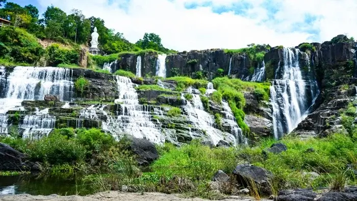 Pongour Waterfall flowing over rocky tiers with a white statue on the cliff above, a natural highlight near Dalat, Vietnam