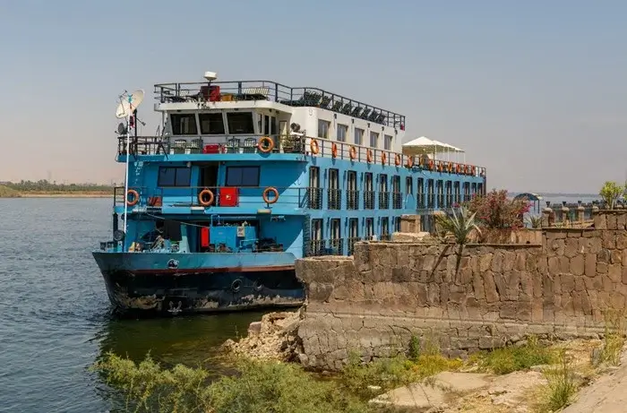 Blue and white cruise boat moored beside an old stone wall on the Nile with orange life rings along its decks
