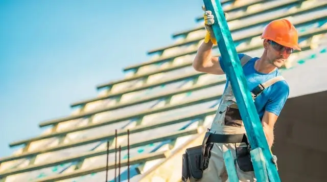 Roofer in a hard hat and safety glasses carrying a wooden batten past an unfinished roof frame with underlayment