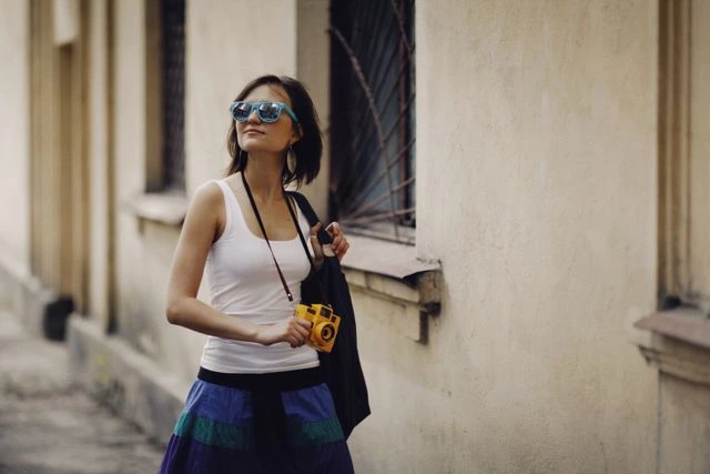 Woman in blue sunglasses holding a yellow camera while exploring an old city street on her own, capturing new travel memories