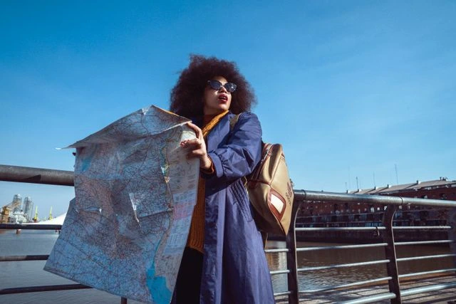 Woman in sunglasses and a navy coat holding an unfolded map by a waterfront railing, finding her own way on a solo trip
