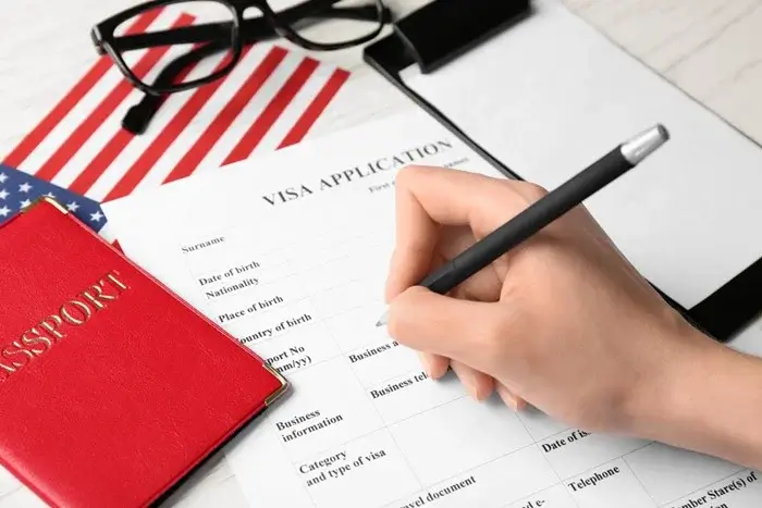 Hand filling out a visa application form with a pen beside a red passport, an American flag and glasses, with an onward ticket needed as proof of travel