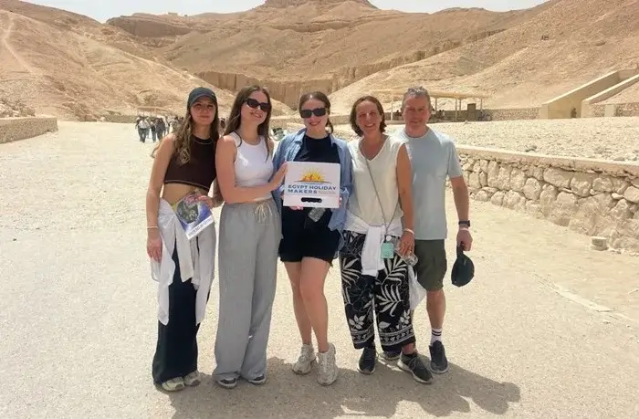 Group of five travelers smiling in the Valley of the Kings in Luxor, holding an Egypt Holiday Makers sign beneath the desert cliffs