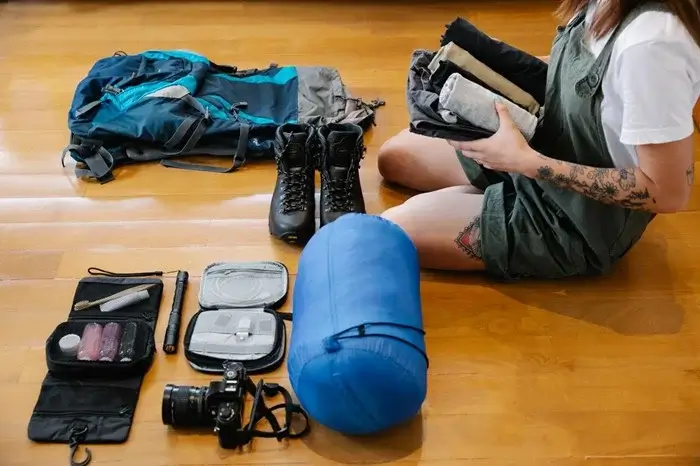 Hiker packing gear on a wooden floor, including a backpack, hiking boots, a camera with a zoom lens, a sleeping bag, and folded layers for a wildlife watching trip
