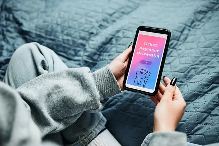 Person sitting on a quilted bed holding a smartphone that shows a “ticket payment successful” screen after booking an onward ticket online