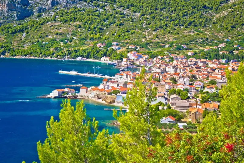 Coastal village on Croatian Dalmatian coast with red-roofed houses, turquoise bay, and forested hills overlooking Adriatic Sea