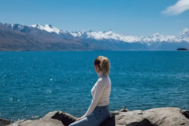 Woman in cream sweater and jeans sitting by mountain lake - preppy travel outfit essentials for scenic destinations