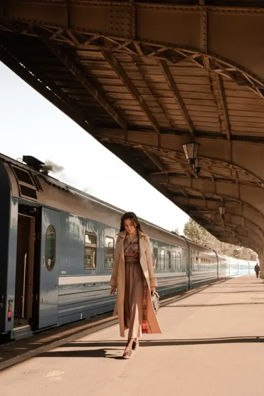 Woman in beige trench coat at vintage train station - classic preppy travel packing essential for European trips