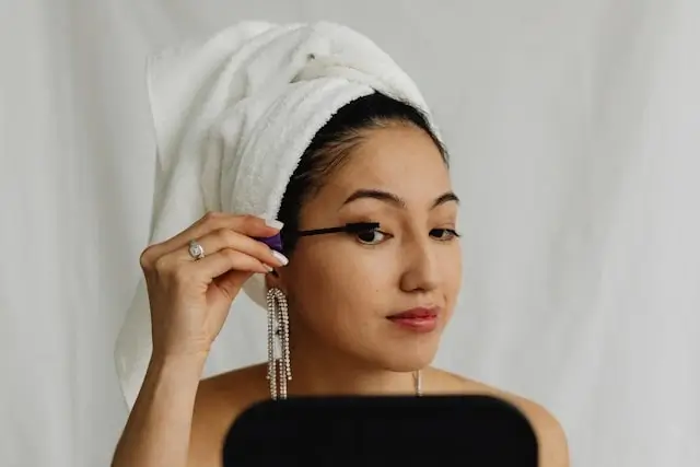 Alt text: "Woman applying mascara to enhance lash growth with white towel wrapped around head during beauty routine