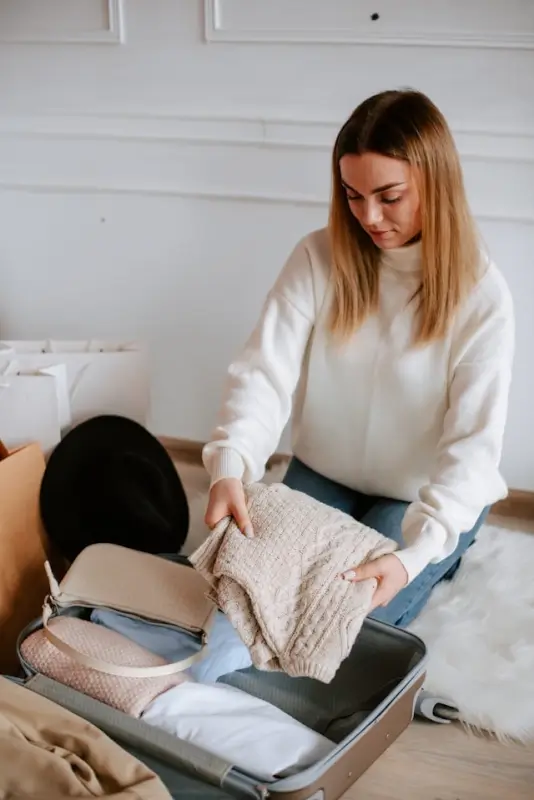 Woman packing cream knit sweater in suitcase - preppy travel essentials for stylish vacation packing.