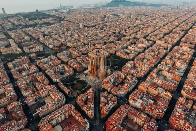 Aerial view of Barcelona's Eixample district showing the distinctive octagonal city blocks arranged in a geometric grid pattern, with the iconic Sagrada Família basilica prominently featured in the center, surrounded by buildings with characteristic reddish-orange tile roofs extending toward the Mediterranean coast in the background.