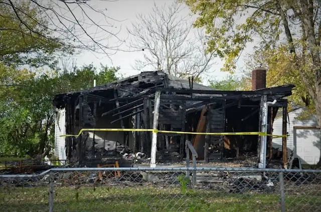 Fire-damaged house with severely burned structure and collapsed roof sections. Yellow caution tape surrounds the property, and a chain-link fence runs along the foreground. The building shows extensive fire damage with blackened walls, exposed framing, and debris scattered around the foundation. Trees with spring foliage frame the scene under an overcast sky.