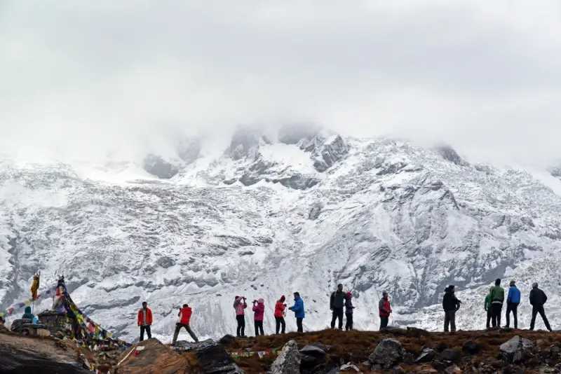 Trekkers in colorful winter gear gather on a rocky viewpoint along the Annapurna Base Camp trail, gazing at snow-covered Annapurna peaks shrouded in clouds, with traditional Tibetan prayer flags in the foreground