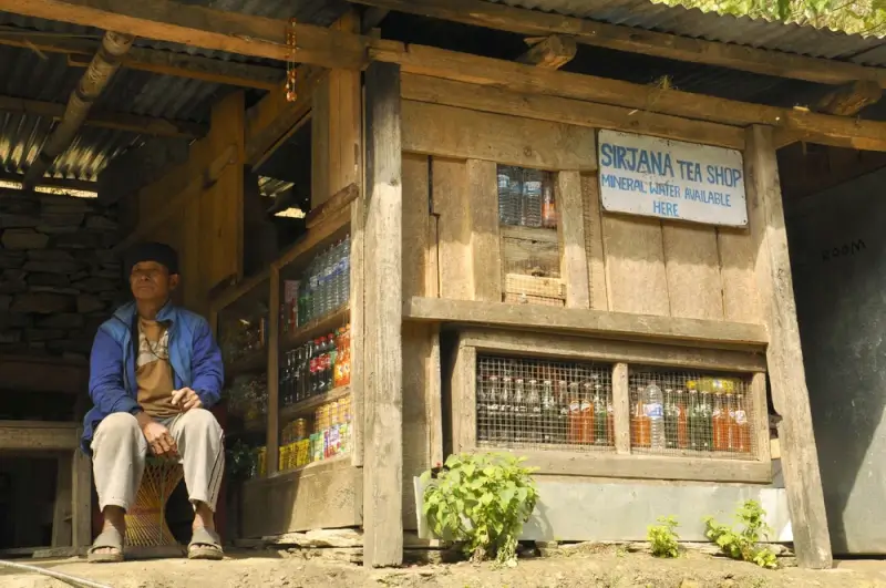 A local man sits outside Sirjana Tea Shop, a traditional tea house along the ABC Trek route offering refreshments and mineral water to trekkers, with goods displayed behind wire mesh windows