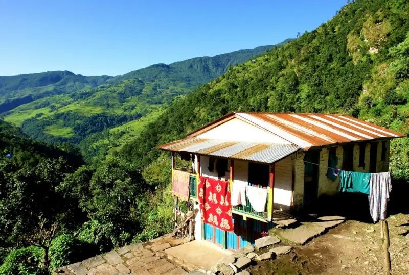 A traditional Nepalese house with a corrugated metal roof sits on a hillside along the ABC Trek, with colorful clothing hanging to dry and lush green terraced hills rolling into the distance under a clear blue sky