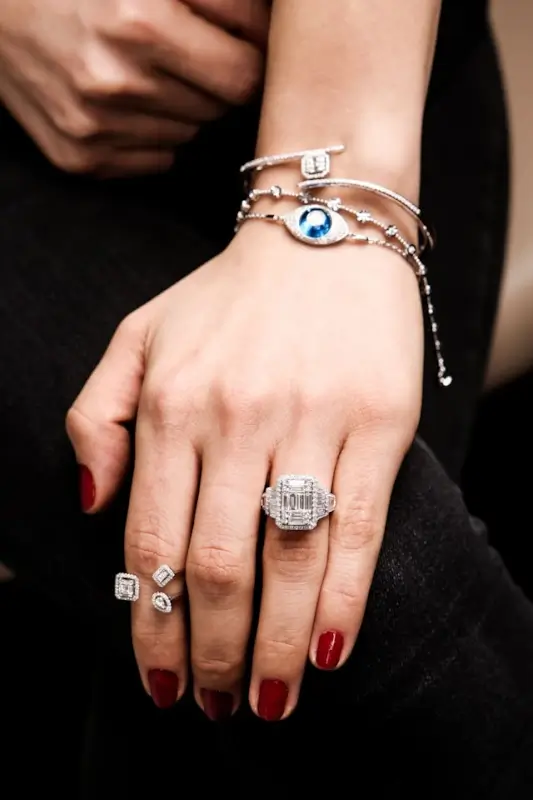 Woman's hand displaying layered gold jewelry including evil eye bracelet with blue stone center and diamond rings with burgundy manicured nails