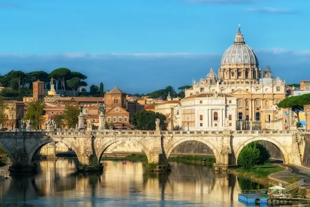 St. Peter's Basilica with its iconic dome dominates the Vatican skyline, viewed across the Tiber River with a historic stone bridge in the foreground during golden hour lighting