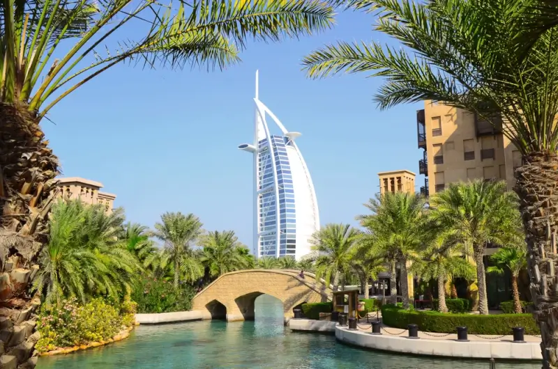 View of Dubai's iconic sail-shaped Burj Al Arab hotel rising above traditional Middle Eastern architecture, framed by lush palm trees and an ornate stone bridge crossing a turquoise waterway in the Madinat Jumeirah resort area.