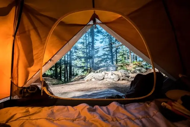 View from inside an orange tent looking out through the open entrance at a forest campsite with tall evergreen trees and rocky terrain. Camping gear and sleeping materials are visible in the foreground inside the tent, creating a cozy outdoor shelter perspective.