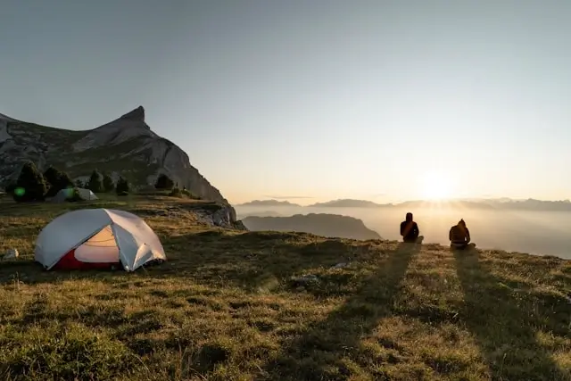 White and red tent positioned on a grassy hillside with two silhouetted figures sitting and watching the sunset over a dramatic mountain landscape. The scene captures golden hour lighting with layered mountain ridges extending into the distance under a gradient sky, creating a serene wilderness camping moment.
