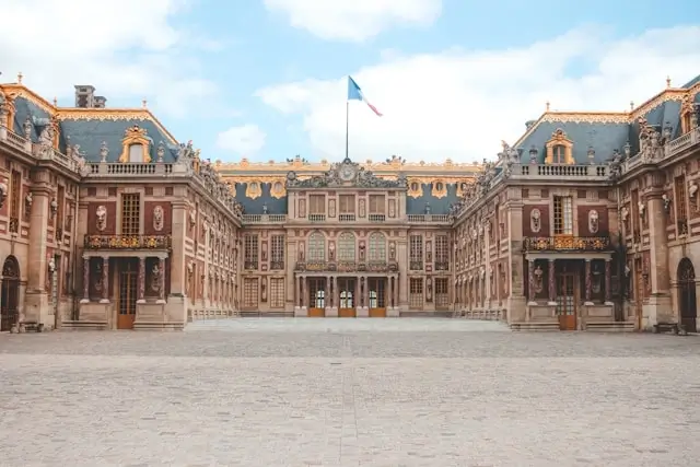 The grand courtyard of Palace of Versailles showcasing symmetrical baroque architecture with ornate golden details, mansard roofs, and a French flag flying above the central entrance