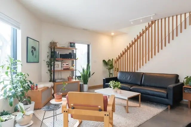 Modern minimalist living room featuring a black leather sofa, wooden coffee table, and prominent wooden staircase with vertical slat railings. The space includes multiple potted plants, built-in shelving with books and decor, a mid-century modern wooden chair, and natural light from large windows. The room has white walls, track lighting, and an area rug, creating a clean aesthetic with warm wood accents.