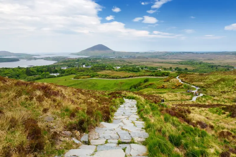 Stone flagstone path winding through Irish moorland with mountain views and scattered white houses in valley