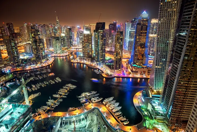 Spectacular nighttime aerial view of Dubai Marina with illuminated skyscrapers reflecting in the water, luxury yachts moored along the curved waterfront promenade, and colorful LED-lit bridges connecting the vibrant entertainment district.
