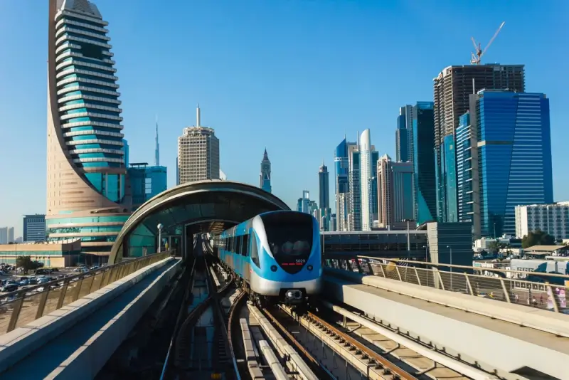 Modern Dubai Metro train with blue and white livery traveling on elevated tracks through the city center, with Dubai's iconic skyline including the Burj Khalifa towering in the background under clear blue skies.