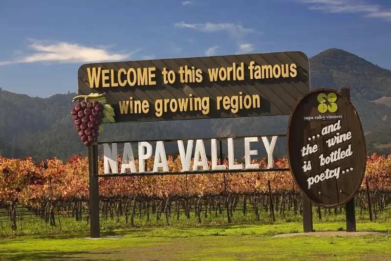 Napa Valley welcome sign reading 'Welcome to this world famous wine growing region' with grape cluster decoration, set against autumn vineyards and rolling hills