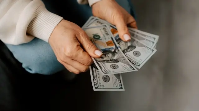 Person's hands counting or handling several one hundred dollar bills. The person is wearing a cream-colored ribbed sweater, and the background is softly blurred. The focus is on the cash being held and examined.
