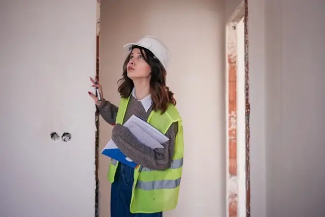 Female construction inspector or building professional wearing a white hard hat and bright yellow safety vest, holding documents while examining an unfinished interior space. She is looking upward, likely inspecting the ceiling or upper walls of a room under construction with exposed electrical outlets and unfinished drywall visible.