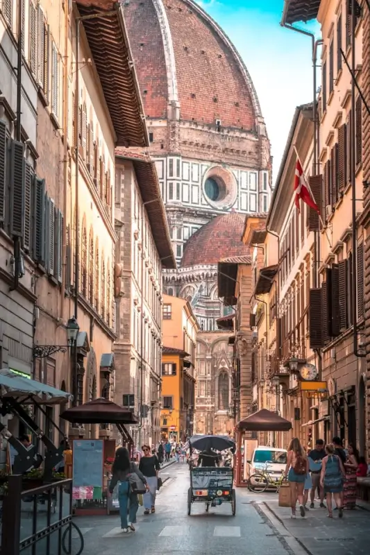 Narrow Florence street leading toward the Duomo cathedral dome, with tourists walking along cobblestones, traditional shuttered buildings, and a small delivery truck navigating the historic pedestrian area