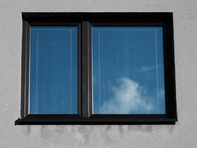 Modern black-framed window with two glass panes set in a gray concrete or stucco wall, reflecting blue sky and white clouds. The window features a clean, contemporary design with dark trim against the neutral exterior wall surface.