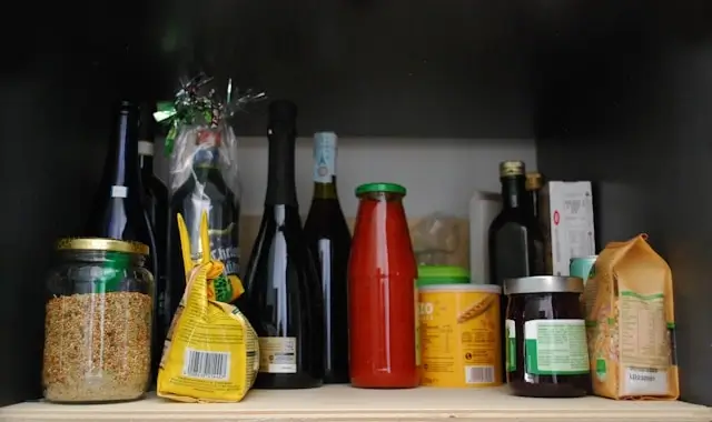 Pantry shelf displaying various food storage containers and ingredients including glass jars with grains or cereals, dark bottles, a red sauce or condiment jar, yellow packaged goods, and a milk or juice carton, all arranged on a wooden shelf against a dark background.
