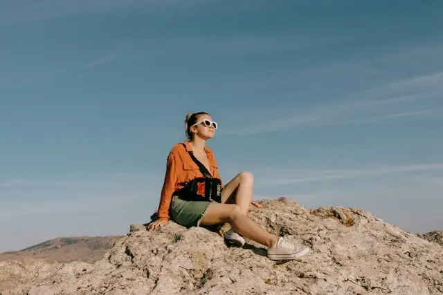 Woman in orange shirt and green shorts with white sunglasses on rocks - colorful preppy travel essentials for desert trips