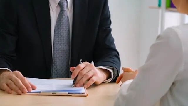 Insurance agent or adjuster in formal business attire reviewing claim documents during a client meeting. The professional is seated at a desk with paperwork, while a client in white is partially visible across the table, depicting a typical insurance consultation scenario.