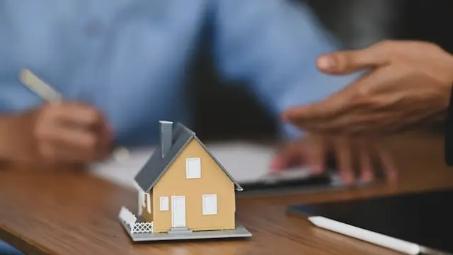 Small yellow house model with gray roof positioned on a white surface in the foreground, with blurred hands of a person in a blue shirt holding papers and working at a desk in the background. The scene suggests a real estate, insurance, or home-related consultation or transaction.