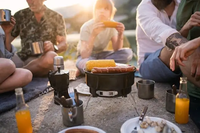 Group of friends gathered around a portable camping grill cooking corn on the cob and sausages during an outdoor camping meal, with camping equipment, coffee maker, metal cups, and orange juice visible on the ground. The scene is lit by warm golden hour sunlight, creating a cozy social camping atmosphere.