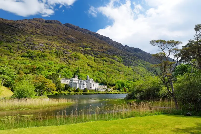 White castle abbey beside tranquil lake with dramatic green mountains and lush Irish countryside
