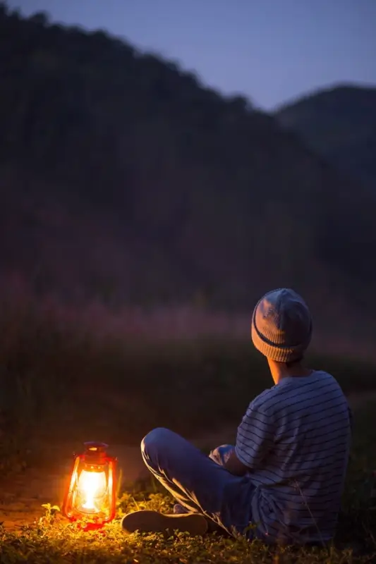 Person in striped shirt and knit beanie sitting cross-legged beside a glowing camping lantern at dusk, with silhouetted mountains in the background. The warm lantern light illuminates the ground around the camper, creating a peaceful, contemplative evening scene in the wilderness.