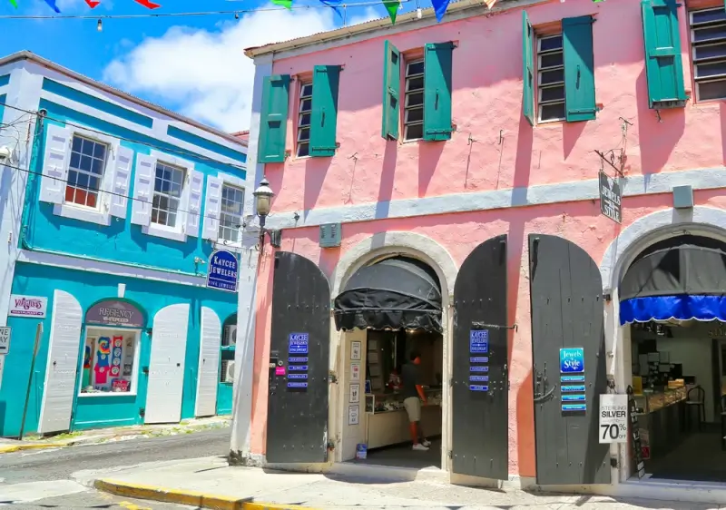 Colorful Caribbean colonial street with turquoise and coral pink buildings featuring green shutters, street-level shops, and festive bunting flags under bright blue sky