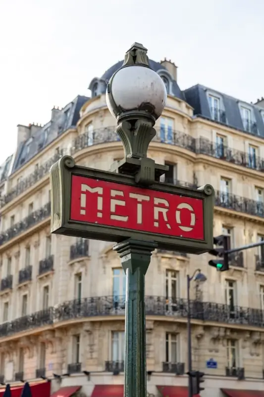 Classic Parisian Metro sign with red lettering and ornate Art Nouveau design, positioned in front of traditional Haussmann-style buildings with wrought iron balconies