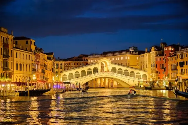 Rialto Bridge illuminated at night over Grand Canal with colorful Venetian palazzos - must-see Venice travel guide destination